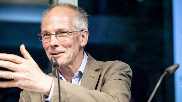 Ein älterer Herr mit Sakko, Brille und schütterem Haar. Er lächelt, hat die Hand gestikulierend erhoben und scheint gerade in ein Mikrofon vor ihm zu sprechen. Ein älterer Herr mit Sakko, Brille und schütterem Haar. Er lächelt, hat die Hand gestikulierend erhoben und scheint gerade in ein Mikrofon vor ihm zu sprechen.